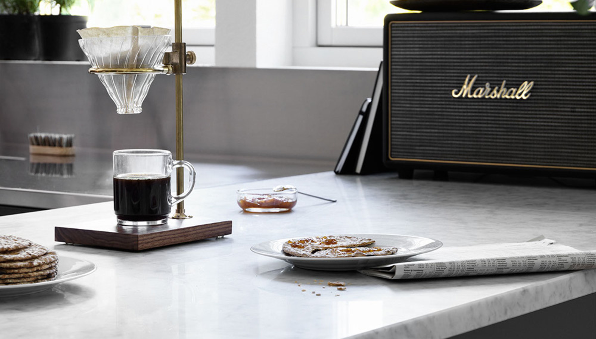 nomenu Manuell kaffebryggare på en marmorbänk med en kopp kaffe bredvid, tillsammans med knäckebröd, sylt och en tidning. En Marshall-högtalare syns i bakgrunden.