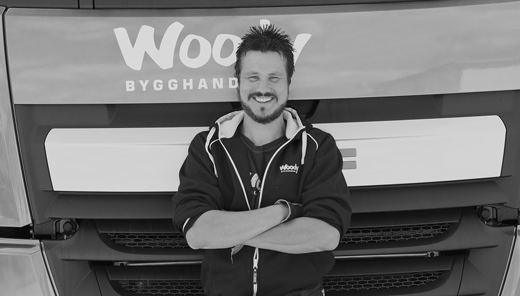 nomenu Säkra materialleveranser, bild på en glad man från Woodys bygghandel som står med armarna i kors framför en lastbil