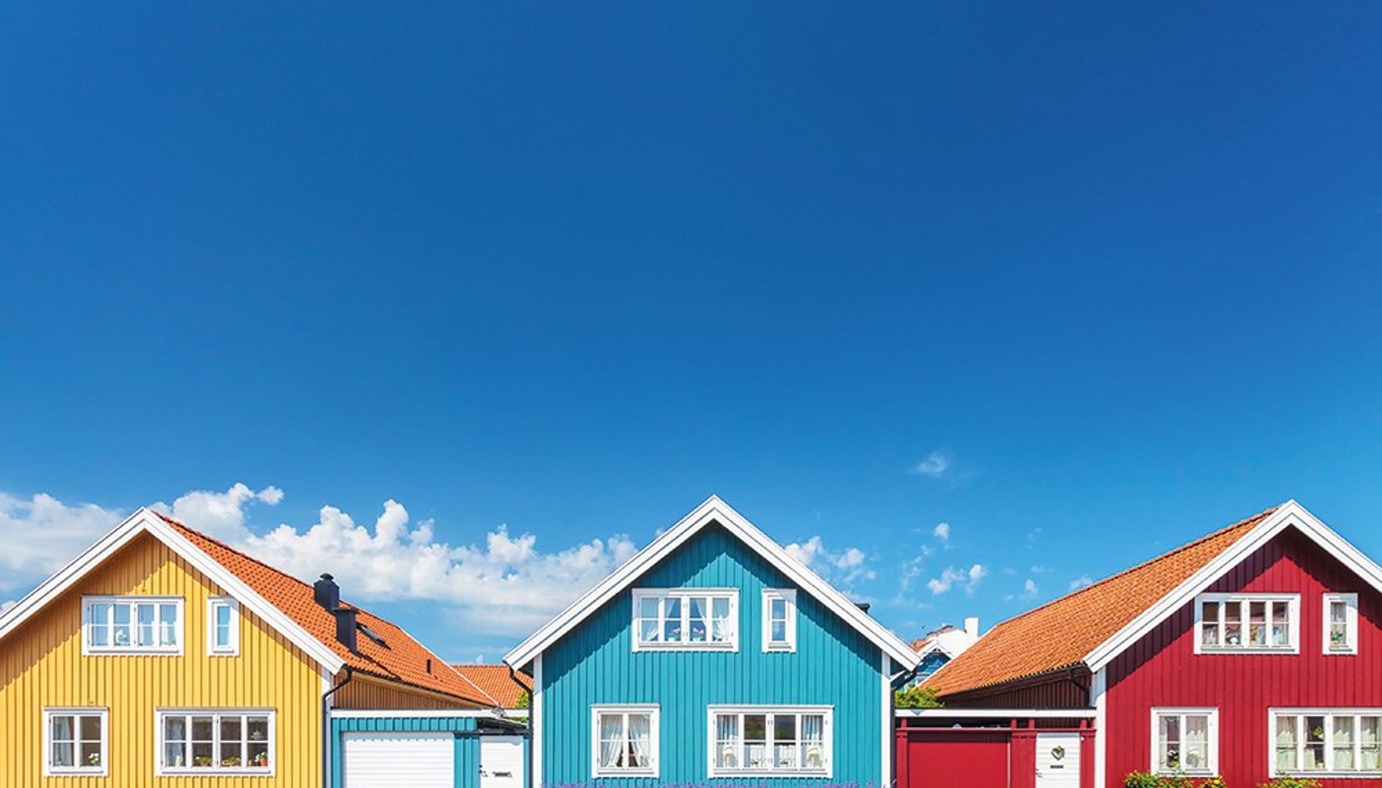 nomenu Bild på tre hus sidan om varandra, ett gult, blått och ett rött hus med många fönster på deras framsidor. 
