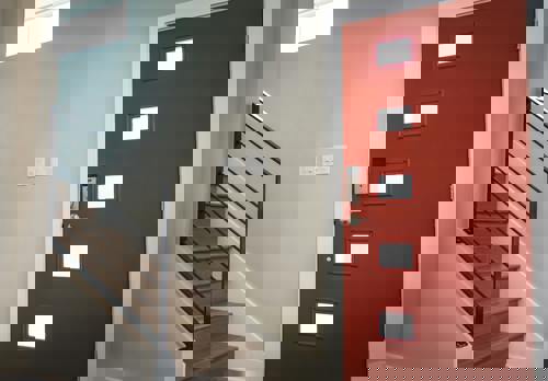 logo Bilden visar en ljus och modern hall med en iögonfallande röd ytterdörr som har fyra små fyrkantiga glasrutor och ett digitalt dörrlås. Bredvid dörren syns en trappa med trästeg och svart metallräcke som leder uppåt
