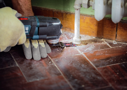 Någon använder en elektrisk slipmaskin på ett golv av mörka plattor nära en vit rörledning.