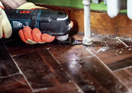 Hand i röda handskar håller elektriskt verktyg mot brunt trägolv bredvid rör och grön vägg.