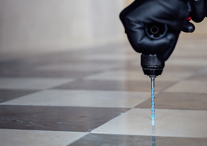 Svartklädd person använder en blå borr på pricklösa, svartvita rutiga plattor.