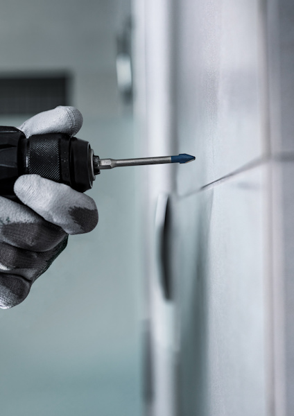 Hand i handske håller en borrmaskin mot kakelvägg.