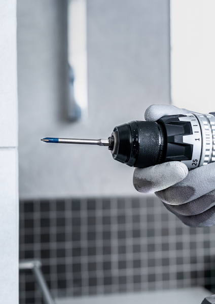 Hand i handske håller borrmaskin nära kakelvägg.