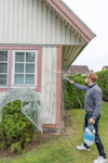Man i trädgård sprayar medel på smutsig husfasad av trä, plast täcker fönster och buske.