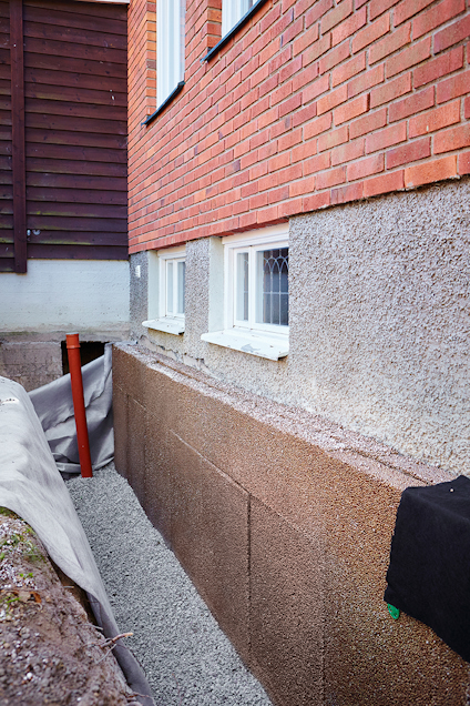 Röd tegelvägg med synlig källargrund och isoleringsskivor intill. Grus längs basen av byggnaden.