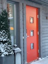 Röd ytterdörr med tre fönsterrutor, omgiven av grå träpanel, brevid en snötäckt buske och en lykta.