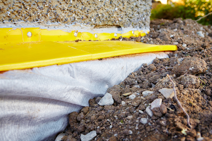 Gul fuktspärr och vit duk installerad vid en grundmur, omgiven av jord och stenar.