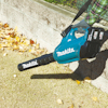 En person använder en blå Makita lövblåsare med batteripaket på en gräsmatta med fallna löv.