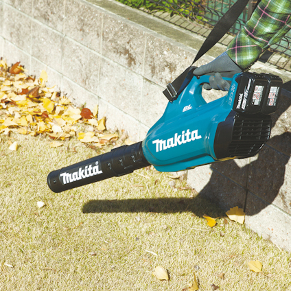 En person använder en blå Makita lövblåsare med batteripaket på en gräsmatta med fallna löv.