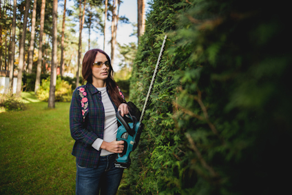 Kvinna med solglasögon används en häcksax för att klippa en stor grön häck i en trädgård.