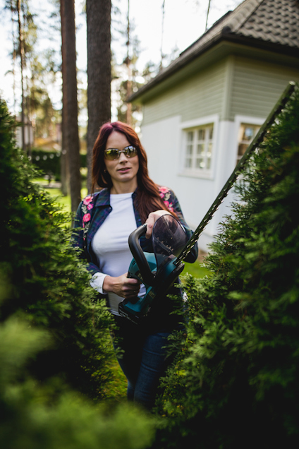 Kvinna med hörselskydd klipper buskar med en batteridriven häcksax nära ett hus.