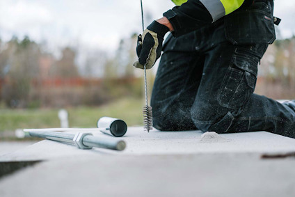 Kneeling worker with high-visibility jacket using a wire brush on concrete, with tools in the background.