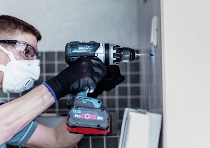 Person med skyddsmask och glasögon borrar i en kaklad vägg med en elektrisk borrmaskin.