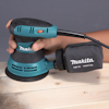 Person använder en blå och svart Makita slipmaskin på träyta.