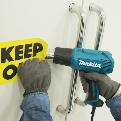 En person håller en blå varmluftspistol nära en gul uppmärkningsskylt med texten "KEEP OUT".