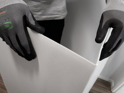 Händer med svarta handskar håller i en vit, flexibel skiva mot en träfärgad golvyta.