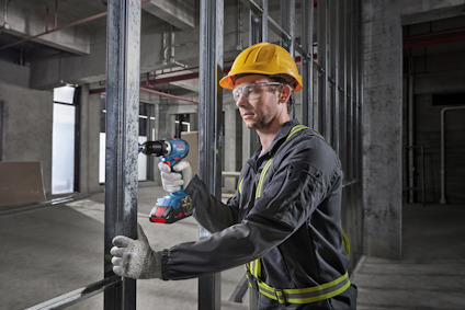 Person med gul hjälm och skyddsglasögon använder en borrmaskin på en byggarbetsplats.