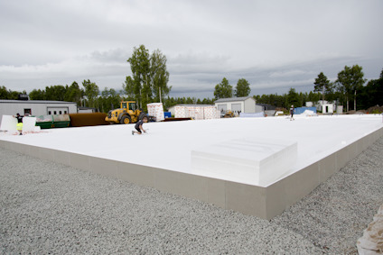 Vit byggplatta på grusunderlag, omgiven av skog och maskiner, med människor som arbetar runt omkring.