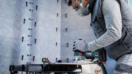Person i skyddsglasögon och ansiktsmask använder elverktyg mot en vägg av betongplattor.