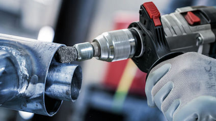 Hand med handske håller en borr som borrar i ett metallrör.