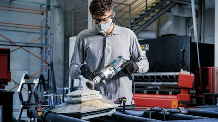 Person med skyddsglasögon och mask slipar något med en elektrisk slipmaskin i en verkstadsmiljö.