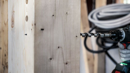 Träskiva med synliga skruvar, en borrmaskin till höger och en rulle kabel i bakgrunden.