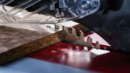 Cirkelsågblad skär genom träplanka, sågspån syns under plankan. Skärande tänder i närbild.