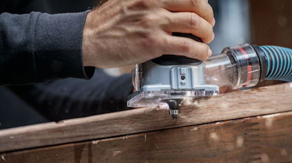 Hand håller en router för träbearbetning över en träyta, ansluten till en dammsugarslang.