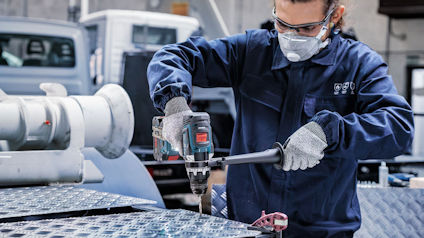 Person i skyddsglasögon och handskar borrar i metallplatta med elverktyg.