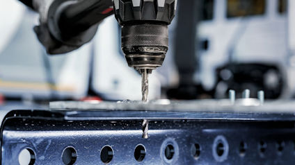 Borrmaskin borrar genom en metallplåt med ett hål i närbild, suddig bakgrund.