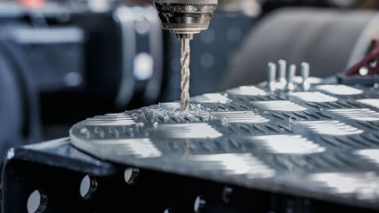 Borrande maskin på en metallplåt med mönstrat yta. Bakgrund suddig verkstadsmiljö.