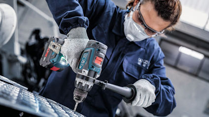 Person med skyddsglasögon och ansiktsmask använder borrmaskin på metallplåt.