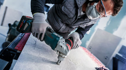 Person som använder handhållen elektrisk borr på en arbetsyta, med skyddsglasögon och mask.