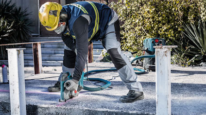 Arbetare med hjälm och skyddskläder använder en vinkelslip för att skära betong utomhus.