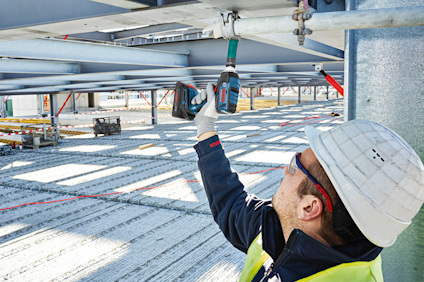 Byggarbetare med skyddshjälm och väst använder elektrisk borr på metallbalk.