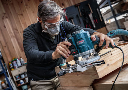 En person i skyddsglasögon och ansiktsmask använder en blå och svart stickfräs på en träbit i en verkstad.