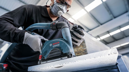 En person slipar med en Bosch-slipmaskin, bär skyddsmask och handskar, i en industrimiljö med lysrör i taket.