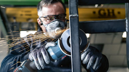 Person slipar metall med vinkelslip, bär skyddsglasögon och ansiktsmask. Gnistregn omkring verktyget.
