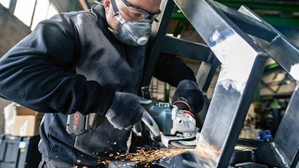 Person i arbetskläder och skyddsutrustning slipar en metallstruktur med vinkelslip, gnistor flyger.