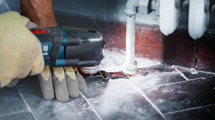 Händer i arbetshandskar använder en elektrisk slipmaskin nära ett rör vid ett golv.