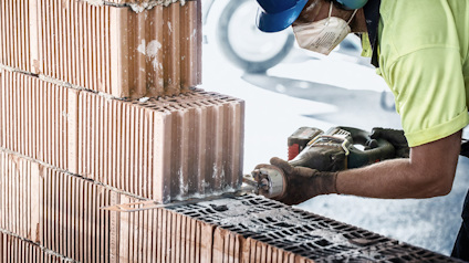 Person med skyddsmask och hjälm skär genom tegelblock med en vinkelslip.