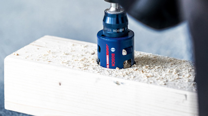 Borra tar sig genom träbit med ett blått hålsågsverktyg, omgiven av träspån.