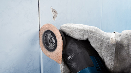 Hand med handske håller en slipmaskin mot en kakelvägg.