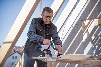En person använder en cirkelsåg på en träskiva, omgiven av trästöd och hus i bakgrunden.