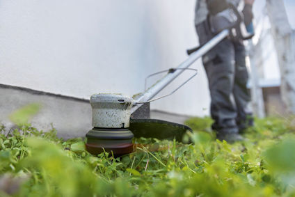 Grästrimmer som används av person i arbetskläder, klipper gräs vid husvägg.