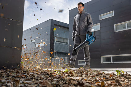 Man i arbetskläder använder en blå lövblåsare för att blåsa bort löv utomhus framför moderna byggnader.