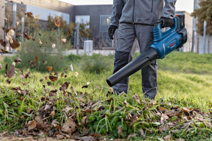 Person i arbetskläder använder en blå lövblåsare för att blåsa bort löv och skräp från en gräsmatta.