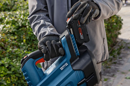 Person med handskar som håller en blå trädgårdsmaskin och sätter i ett batteri märkt "18V 5.0Ah".